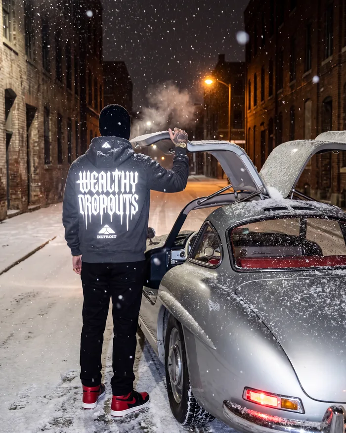 Charcoal heather, worn from behind in falling snow beside a silver Mercedes-Benz 300 SL with its doors raised, the back print lit white by the flash
