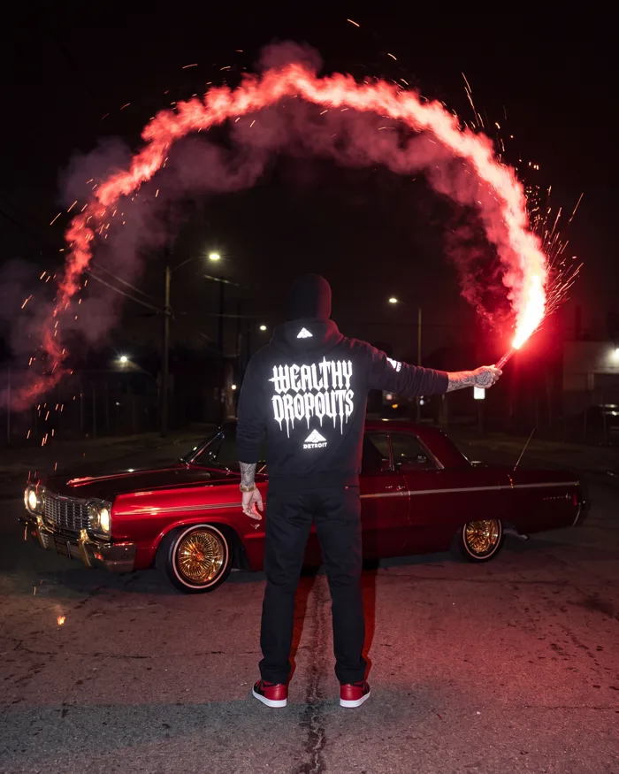 From behind: a man in the black zip hoodie holds a red flare that has traced an arc across a night street, the back print lit white by the flash